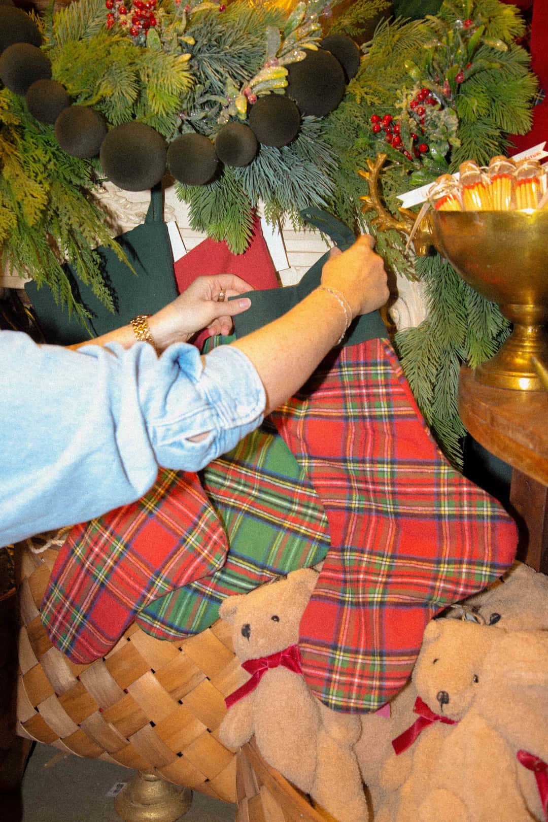 A woman's arms hanging a traditional red and green plaid stocking above a decorated mantle. 
